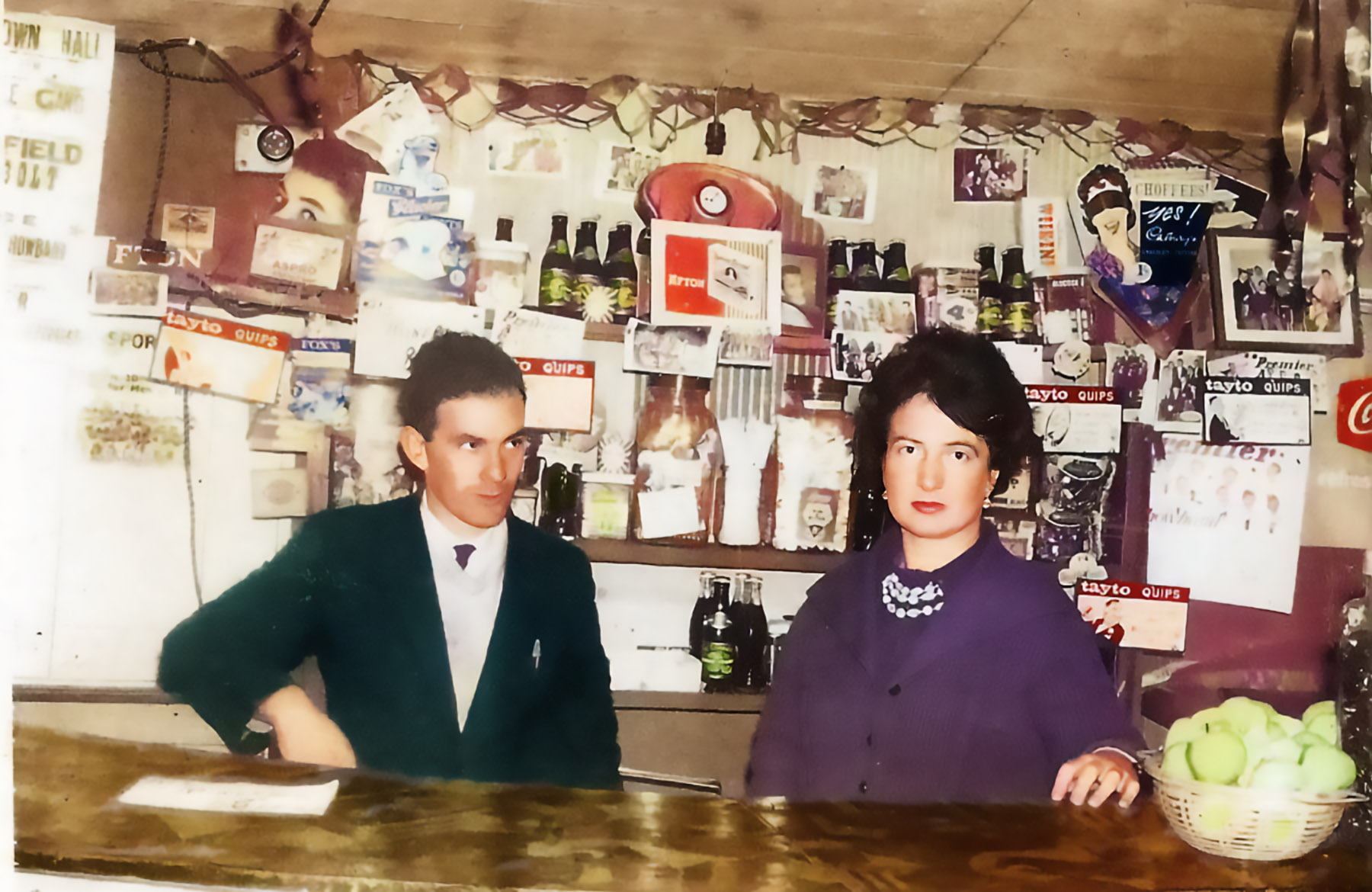 Photograph of Margie Murphy behind the mineral bar which she ran for decades in The Town Hall, Athenry.  She’s accompanied by Tom Fahy who also ran shops in the town including Fahy’s VG Foodmarket on Northgate Street.  Behind them are ads for Fox’s Glacier mints, Afton Cigarettes, Aspro, Cadbury’s Choffees, some Tayto Quips cards, posters for several showbands including the Athenry band Premier Aces. 